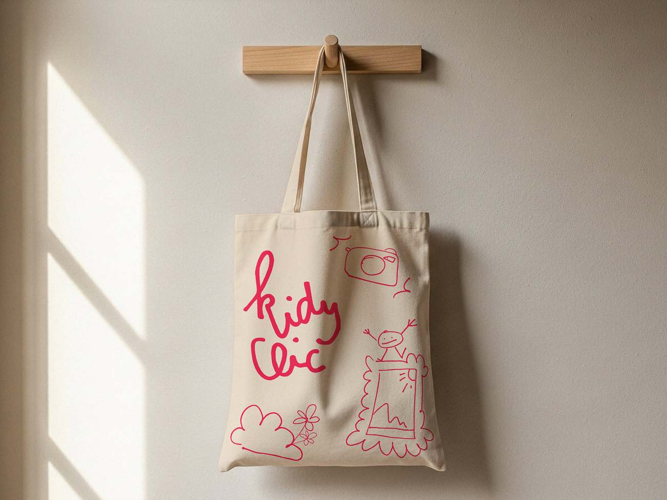 Un tote bag écru accroché au mur, le logo et les dessins en rose vif