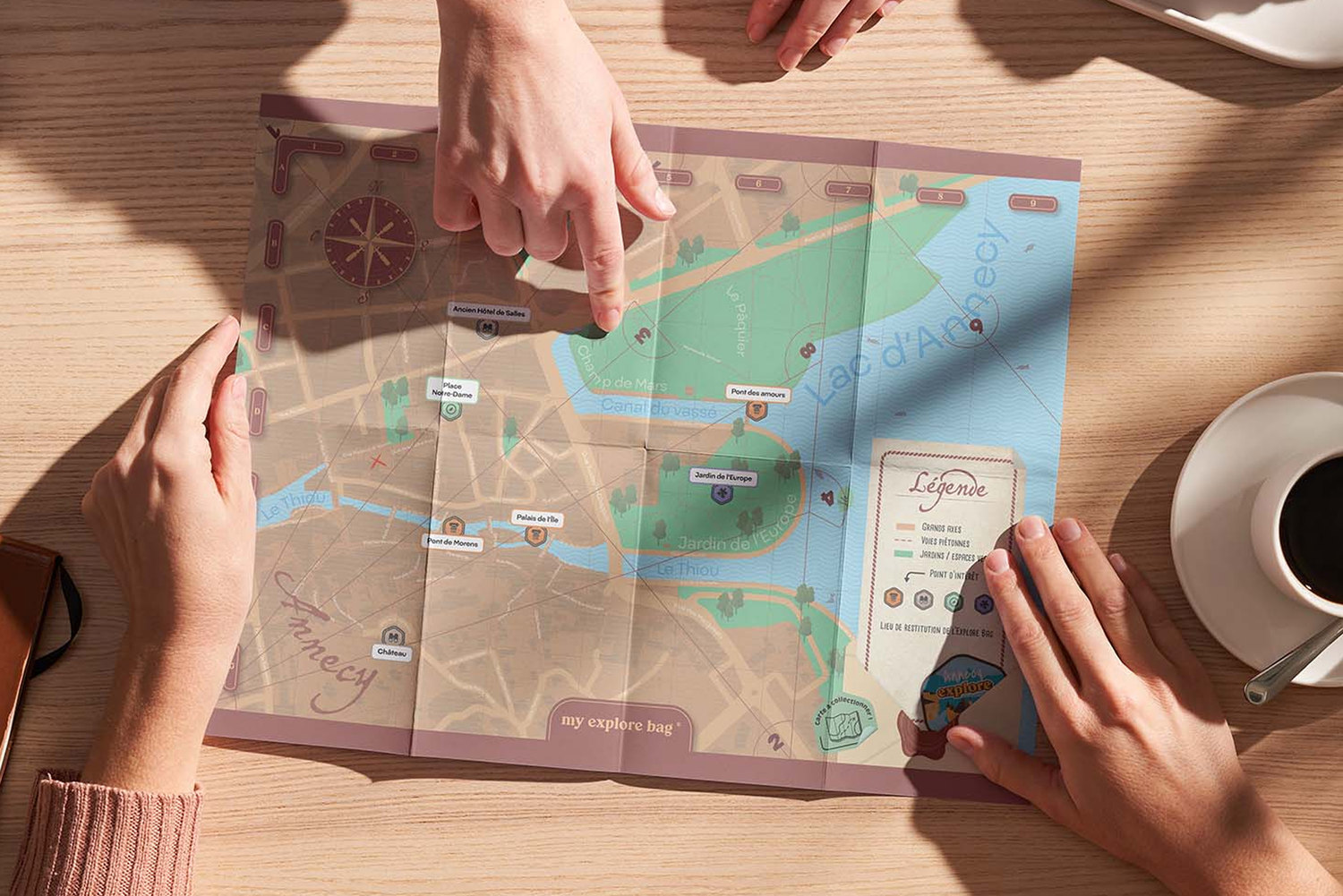 Des mains déplient la carte d’Annecy sur une table, à côté d’une tasse de café
