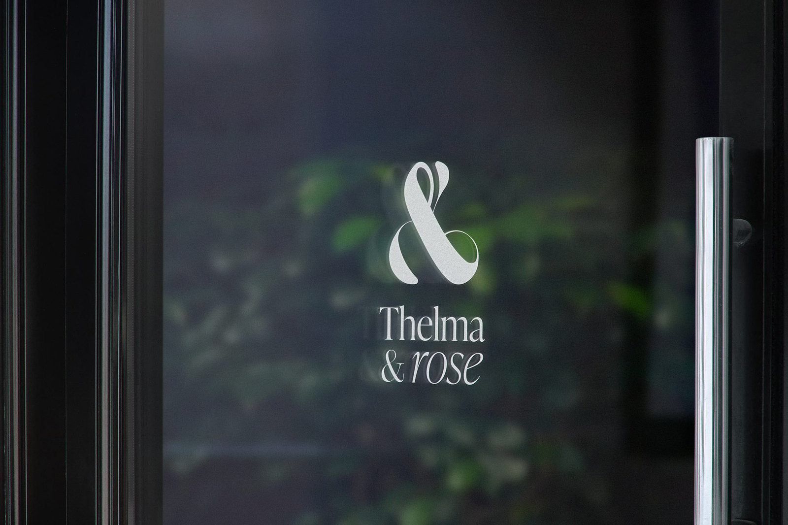 Le logo Thelma & Rose en blanc sur la porte vitrée d’une boutique, de la verdure en reflet